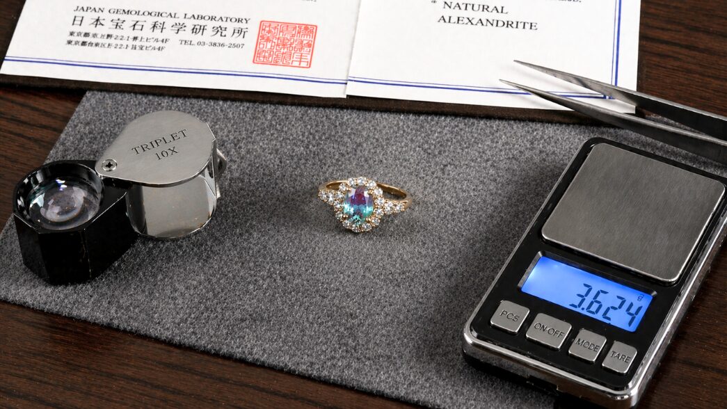 アレキサンドライトリングの横に、鑑定用ルーペ・精密秤・鑑別書が並ぶ「総合評価」の鑑定風景