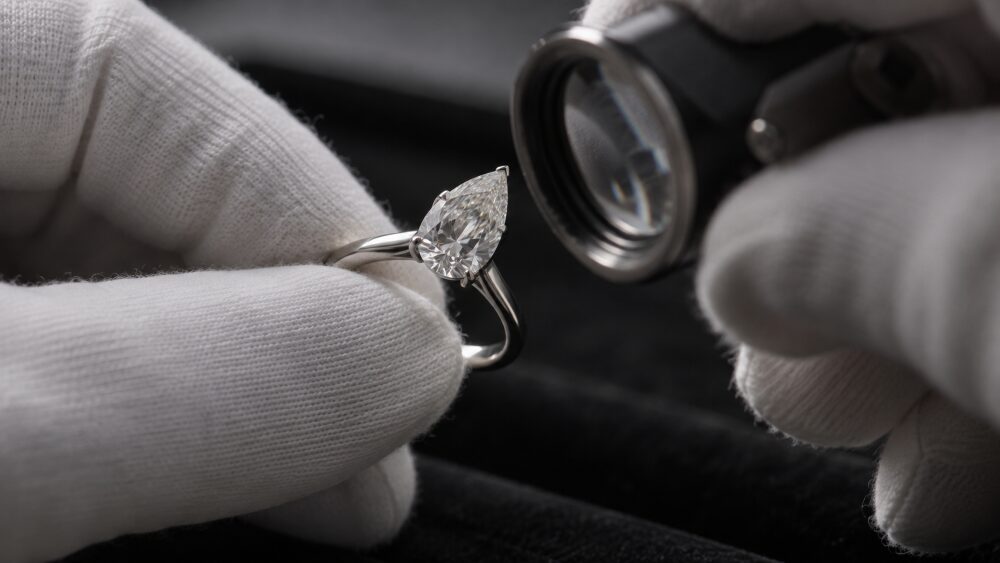 白い手袋をした手がピンセットでしずく型のダイヤモンドの指輪をつまみ、もう一方の手のルーペ越しに石の先端と爪を確かめている手元