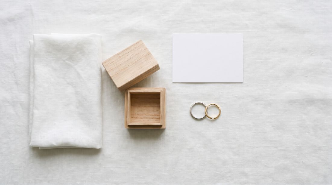 白い麻布の上に並べた結婚指輪2本と、柔らかい布・木箱・書類のイメージ