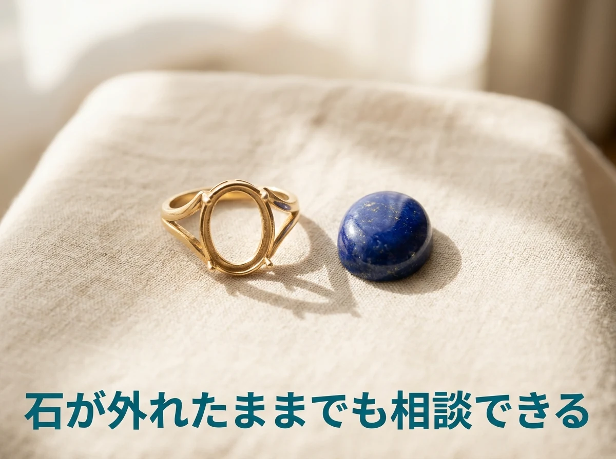 やわらかいリネンの上に、石が外れた金色の指輪の枠と、外れた濃い青のラピスラズリの楕円形の石を並べて置いた写真。石が外れたままでも相談できることを示している。画像内の文字は「石が外れたままでも相談できる」